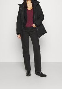 Schwarze Daunenjacke mit Kapuze, bordeauxfarbenes V-Ausschnitt-Shirt, schwarze Jeans und schwarze Stiefel mit Absatz. Einfaches Design und typische Stofftexturen sichtbar.