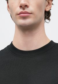 Spodná časť tváre a krku osoby oblečenej v čiernom tričku s okrúhlym výstrihom na pozadí bez vzoru.