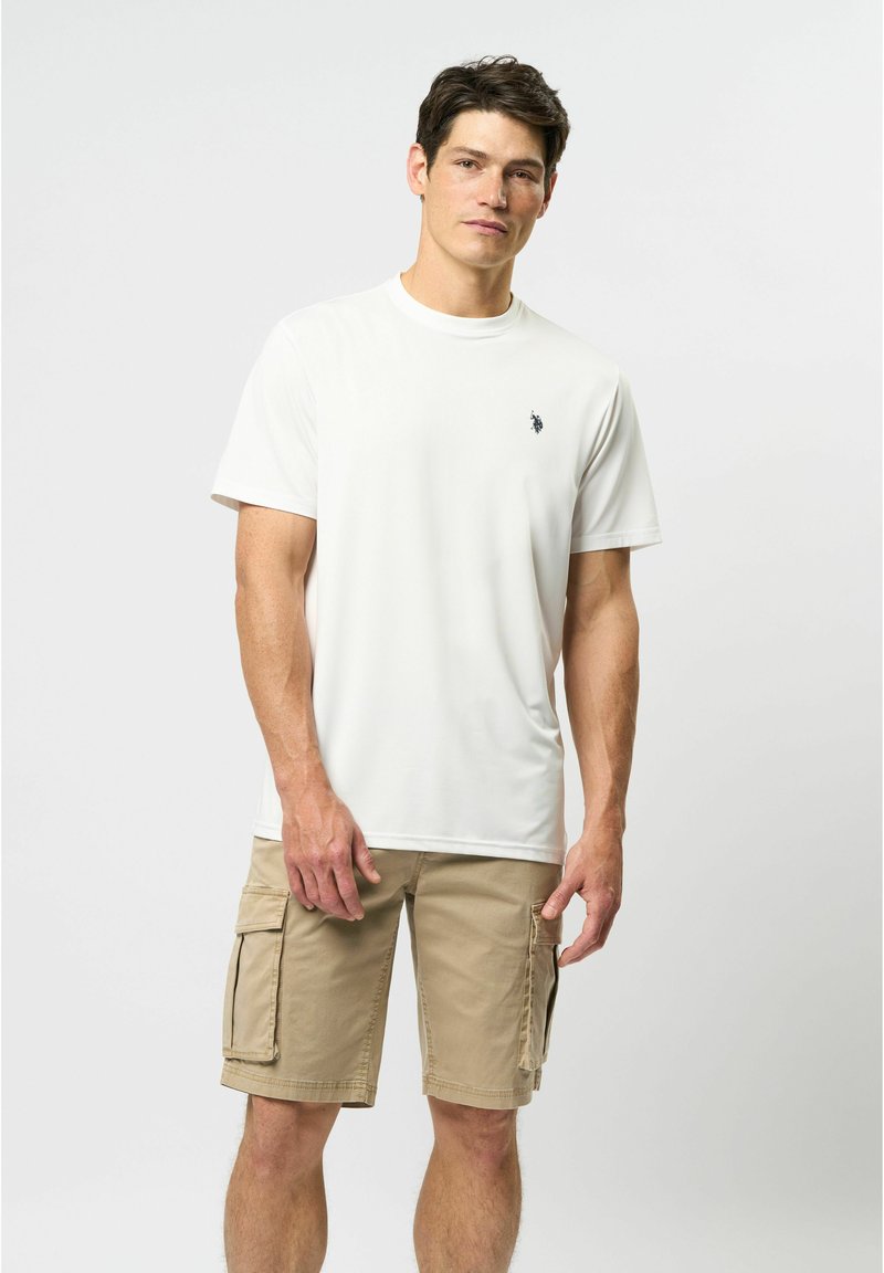 Balta trumpa rankovė t-shirt, pagaminta iš glotnaus audinio, su mažu logo ant krūtinės. Derinama su beige spalvos cargo šortais su šoninėmis kišenėmis.