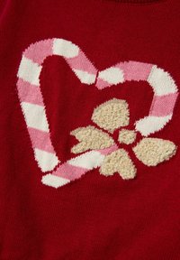 Bastoncino di zucchero a forma di cuore, rosa e bianco a righe, con un fiore beige testurizzato su uno sfondo di tessuto rosso lavorato a maglia.