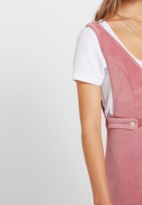 Roze corduroy bolderok met een V-halsontwerp, voorzien van knopen aan de zijkant en een witte geribde t-shirt eronder. Soepele textuur.