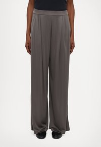 Pantaloni grigi a gamba larga realizzati in tessuto liscia, con pieghe frontali e vita elasticizzata. Abbinati a scarpe nere.