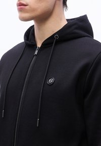 Gros plan sur une personne portant un sweat à capuche noir à fermeture éclair avec des cordons et un petit logo circulaire sur le côté gauche de la poitrine.