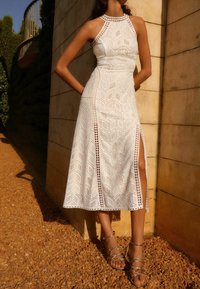 Vestido midi blanco con patrones geométricos, aberturas laterales y diseño de cuello halter. Combinado con sandalias metálicas de tiras.