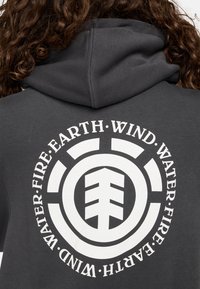 Sudadera gris con un gran gráfico circular en blanco en la parte posterior que presenta palabras elementales: Tierra, Aire, Fuego, Agua, y un icono de árbol en el centro.
