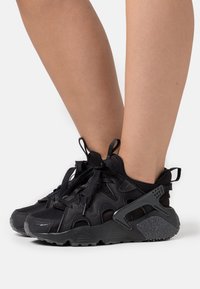 Black athletic sneakers feature a mesh and suede upper, unique cut-out design, textured sole, and laces with a looped pull tab at the back.