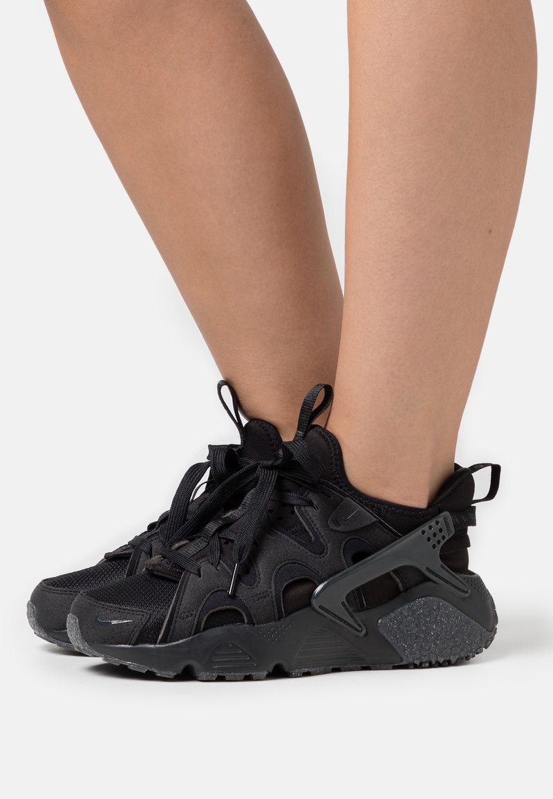Black athletic sneakers feature a mesh and suede upper, unique cut-out design, textured sole, and laces with a looped pull tab at the back.