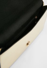 Beige kabelka otevřená, aby ukázala černý interiér s malou zipovou kapsou a zlatým magnetickým zapínáním na přední straně.