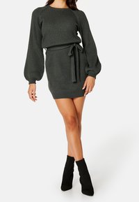 Femme portant une robe en tricot côtelé vert foncé avec des manches bouffantes, une ceinture nouée à la taille, et des bottines noires, debout devant un fond blanc.