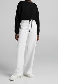 Schwarzer, langärmeliger Pullover mit lockerer Passform, kombiniert mit weißen, hoch taillierten, weiten Jeans. Enthält eine Kordelzugdetail an der Taille.