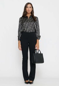 Donna in piedi indossa pantaloni neri e una blusa nera a fantasia, tiene una borsa nera nella mano destra davanti a uno sfondo bianco semplice.