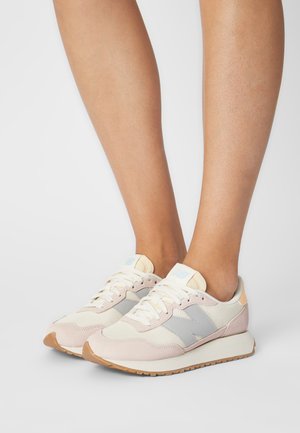 Para beżowych i jasnoszarych sneakersów New Balance noszonych na bose stopy na prostym białym tle.