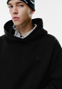 Czarny oversize hoodie z dużym kołnierzem i tonalnym haftowanym logo. Pod spodem widoczna jest koszula w kratę w odcieniach niebieskiego i beżowego.