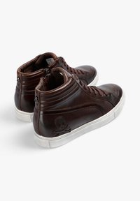 Zapatillas altas de cuero marrón con suelas blancas, que presentan un diseño de calavera y huesos cruzados en el lateral y un cierre de cremallera.