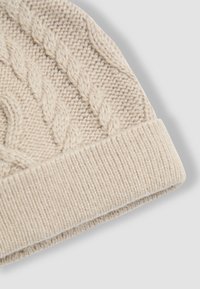Berretto beige lavorato a maglia con motivo a treccia e bordo a coste ripiegato, mostrato su uno sfondo grigio chiaro.