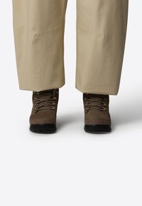 Scarponi da trekking marroni con tomaia in suede texture, occhielli in metallo e suole in gomma nera, abbinati a pantaloni beige a vestibilità morbida.