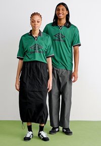 Camisas desportivas verdes com detalhes pretos, combinadas com uma saia preta e calças cinzentas. Ambos usam ténis pretos. Tecido leve, design casual.