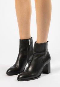 Bottines en cuir noir avec talons carrés et zips latéraux portées sur des jambes nues sur fond blanc.