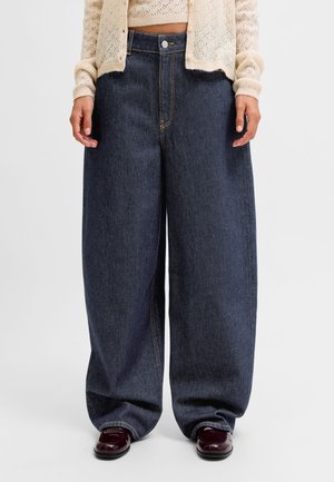 Personne portant un jean large bleu foncé associé à des chaussures bordeaux et un cardigan court en tricot crème, debout sur un fond blanc.