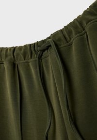 Olivgrüner Jogginghosenbund mit elastischem und verstellbarem Kordelzug. Glatter Stoff mit seitlichen Falten, der eine weiche Textur und eine lockere Passform bietet.