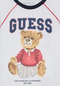 Camiseta blanca con un gráfico de un oso de peluche con una camiseta roja y una falda blanca, con "GUESS" en letras marinas arriba. Textura de algodón suave.