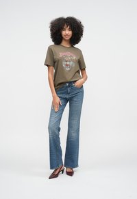 Camiseta gráfica verde oliva con diseño de león, combinada con pantalones vaqueros azules acampanados. El modelo lleva zapatos de tacón granate, posando frente a un fondo liso.