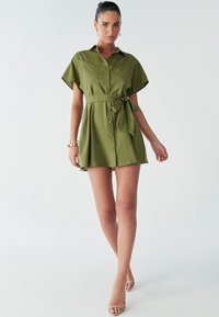 Abito camicia verde khaki con maniche corte, chiusura a bottoni sul davanti, cintura da annodare in vita e vestibilità comoda, realizzato in un tessuto morbido.