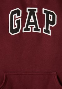 Bordeaux hoodie met een groot, geborduurd "GAP"-logo in zwart met een witte rand, zachte textuur en een voorkantzak.