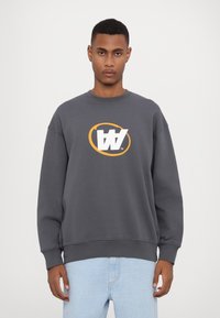 Grijze sweatshirt met een ronde halslijn, voorzien van een opvallend wit en oranje logo op de borst. Zacht materiaal en een losse pasvorm.