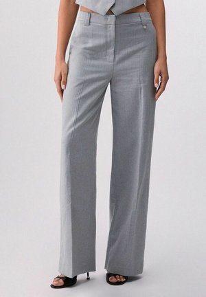 Donna che indossa pantaloni grigi a vita alta e gamba larga, abbinati a tacchi neri aperti sulle dita, in piedi contro uno sfondo chiaro e uniforme.