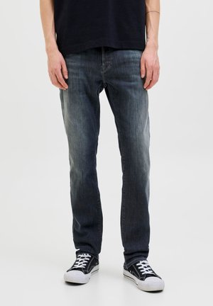 Personne portant un jean bleu foncé délavé et des baskets noires avec des lacets blancs, debout devant un fond uni.