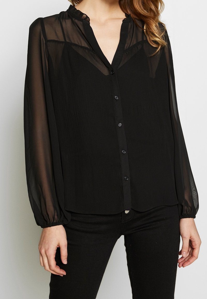 Musta napillinen paita, jossa on poimuttunut rakenne, läpikuultavat pitkät hihat ja röyhelökaulus. Paidan malli on normaali ja siinä on hienovarainen kiilto.