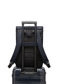 Sac à dos noir et marine avec un design rembourré et des bretelles réglables, reposant sur une valise à roulettes brillante de couleur marine avec une poignée télescopique.