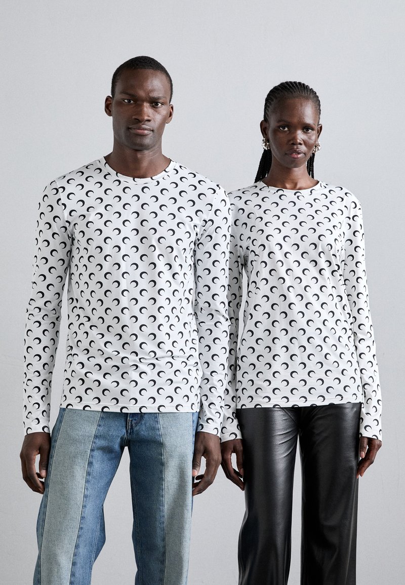 Vit långärmad skjorta med ett mönster av svarta halvmånar. En modell har på sig denimbyxor; den andra har svarta läderbyxor.