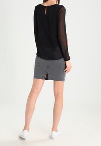 Blusa nera a maniche lunghe in tessuto trasparente con apertura a chiave sul retro. Gonna di jeans grigia con spacco centrale. Sneakers bianche completano l'outfit.