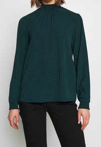 Donna che indossa una blusa a maniche lunghe verde e nera con un colletto alto e arricciato e pantaloni neri, in piedi contro uno sfondo semplice.