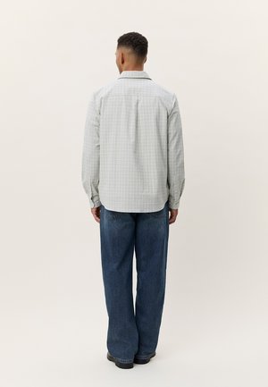 Άνδρας με την πλάτη, φορώντας ανοιχτό γκρι καρό πουκάμισο με μακριά μανίκια και φαρδιά σκούρα μπλε τζιν, στέκεται μπροστά σε ανοιχτό ουδέτερο φόντο.