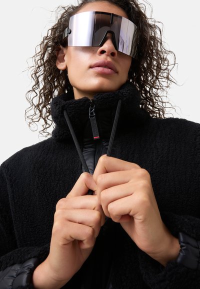 Black fleece jacket with a soft texture, high collar, and a zip closure. Model wears large mirrored sunglasses, enhancing modern aesthetic.