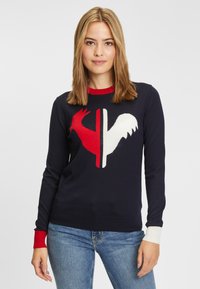 Granatowy sweter z czerwono-białym wzorem koguta, z żeberkowym dekoltem, a także kontrastującymi czerwonymi i białymi ściągaczami. Wykonany z miękkiego materiału dzianinowego.