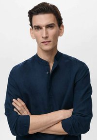 Navy blue linen shirt with a stand collar, button detailing, long sleeves, and a smooth texture. Model stands with arms crossed.