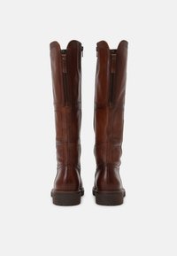 Une paire de bottes en cuir marron hautes vues de l'arrière, avec des fermetures éclair latérales et des détails cousus, posées sur un fond gris clair.