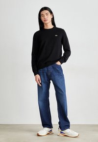 Sort sweater med rund halsudskæring og grønt logo, parret med lyseblå jeans og hvide sneakers med blå og gule accenter.