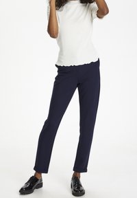 Top bianco a maniche corte con costine e orlo a scallop, abbinato a pantaloni blu scuro. La modella indossa scarpe nere con lacci e finitura liscia.