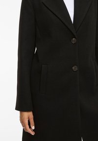 Cappotto nero con design su misura, colletto a revers, due bottoni frontali e tasche laterali; realizzato in un tessuto testurizzato.