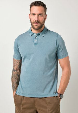 Mann mit tätowiertem linkem Arm, der ein hellblaues Poloshirt und braune Hose trägt, mit Händen in den Taschen vor einfarbigem Hintergrund stehend.
