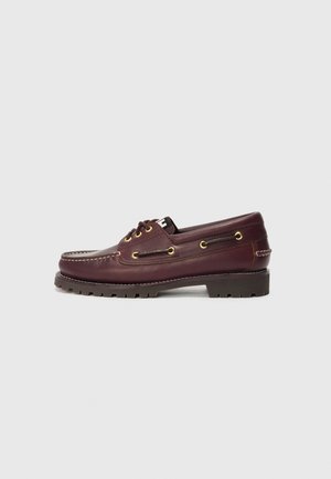 Scarpa da barca in pelle bordeaux con punta arrotondata, superficie testurizzata, occhielli dorati e suola nera robusta. Due lacci decorativi sulla parte superiore.