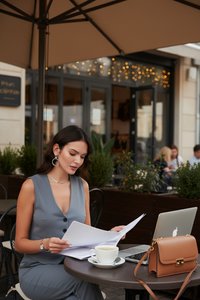 Vrouw zit aan een tafel bij een café buiten documenten te bekijken met een laptop, koffiekopje en handtas onder een grote parasol.
