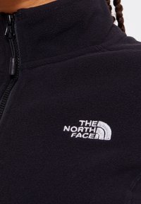 Giacca in fleece nera con colletto alto, dotata di chiusura con zip e un logo ricamato bianco sul petto. Tessuto morbido e strutturato.