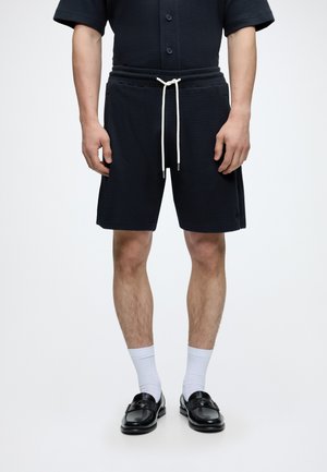 Man draagt zwarte gestructureerde shorts met een witte trekkoord, witte sokken, zwarte leren loafers en een zwart kortärmelig overhemd met knopen.
