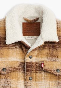 Chaqueta de cuadros en tonos amarillos y marrones, con un cuello de forro polar blanco, botones a presión y una etiqueta de cuero con los detalles de la marca.